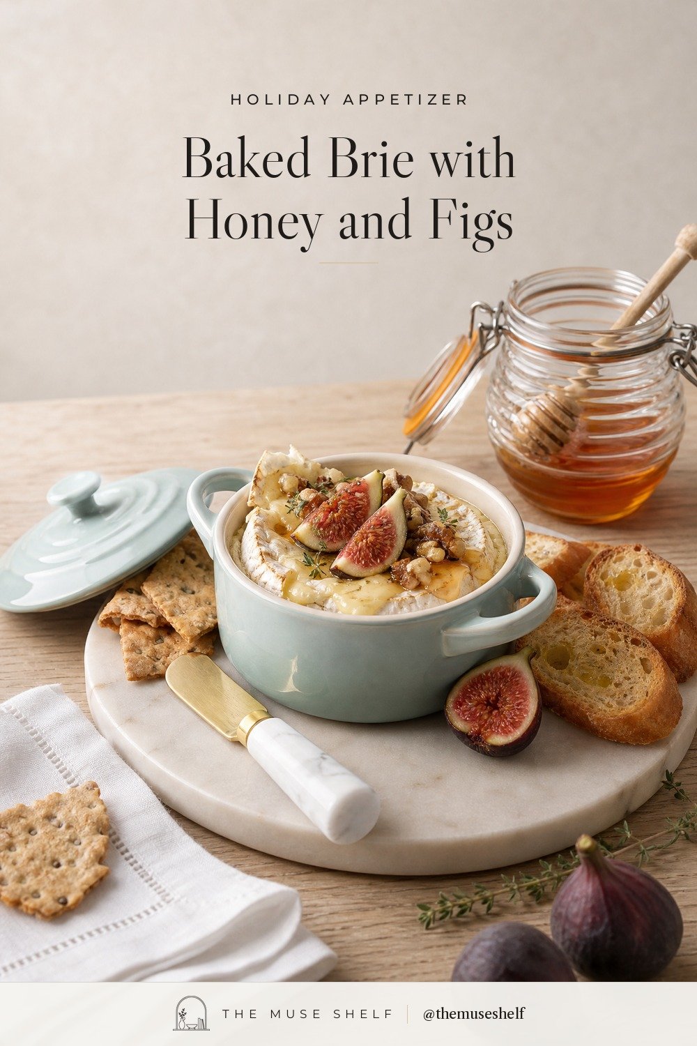 Baked brie with honey, fresh figs, walnuts and thyme in a pale blue-green Le Creuset mini cocotte on a round white marble board, with crackers, toasted baguette, a glass honey pot and a white linen cocktail napkin
