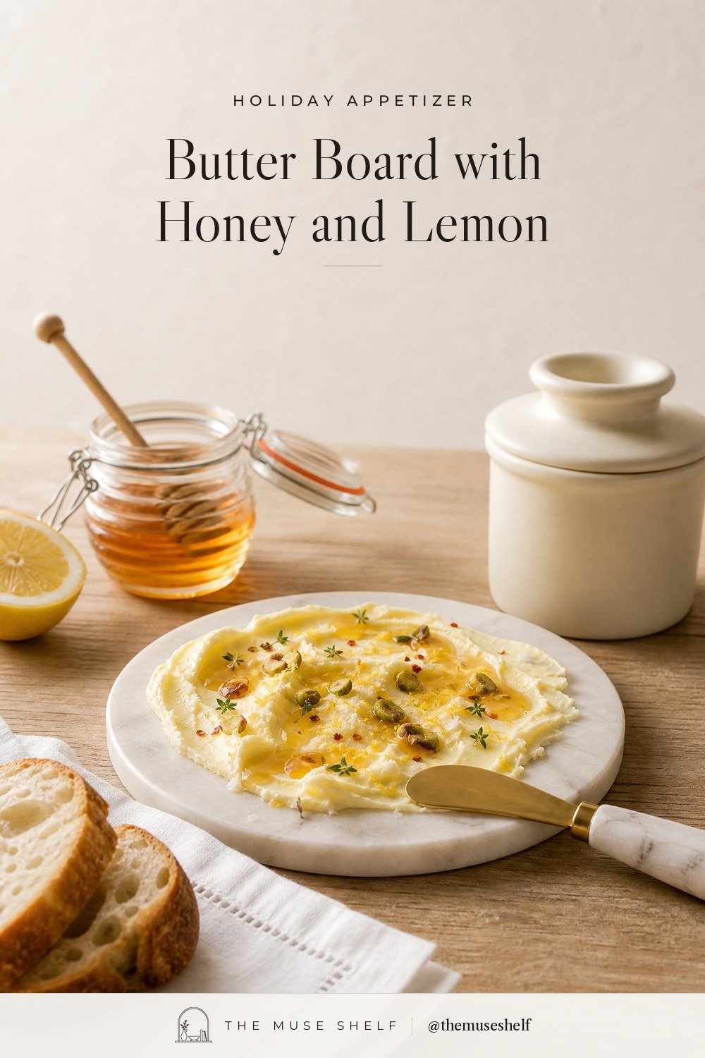 Small butter board on a round white marble board: soft butter swoops with honey, lemon zest, pistachios, thyme and chili flakes, a gold spreader with a marble handle, an ivory Butter Bell crock, a glass honey pot and sliced baguette on a linen napkin