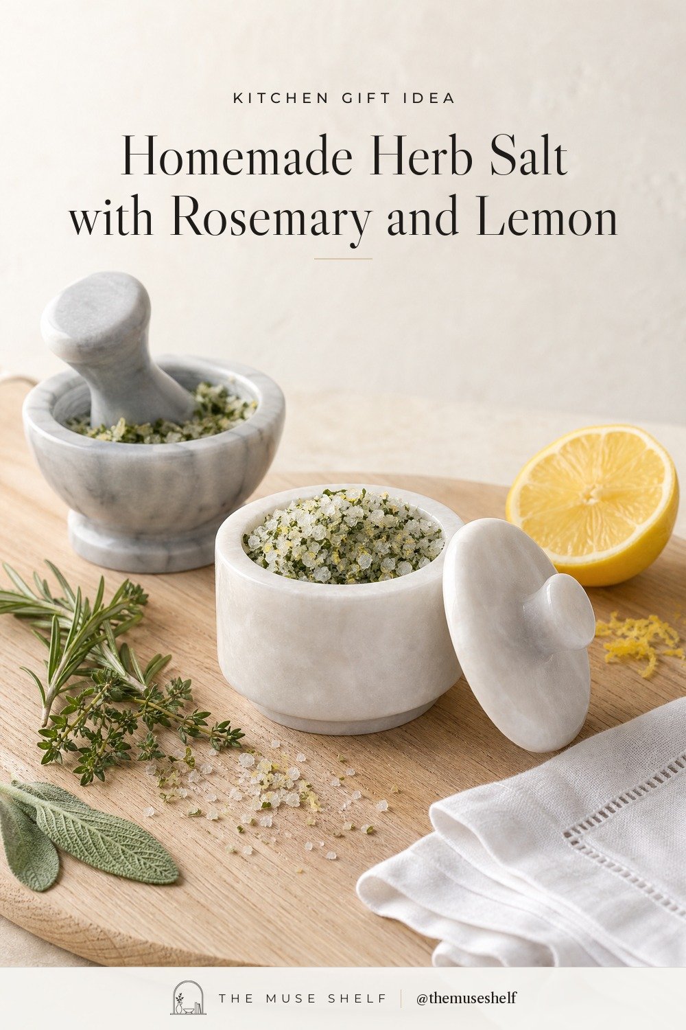 White marble salt cellar filled with coarse green herb salt, its lid beside it, a small marble mortar and pestle with pounded herbs and salt, fresh rosemary, thyme and sage, half a lemon and a linen napkin on an oak board