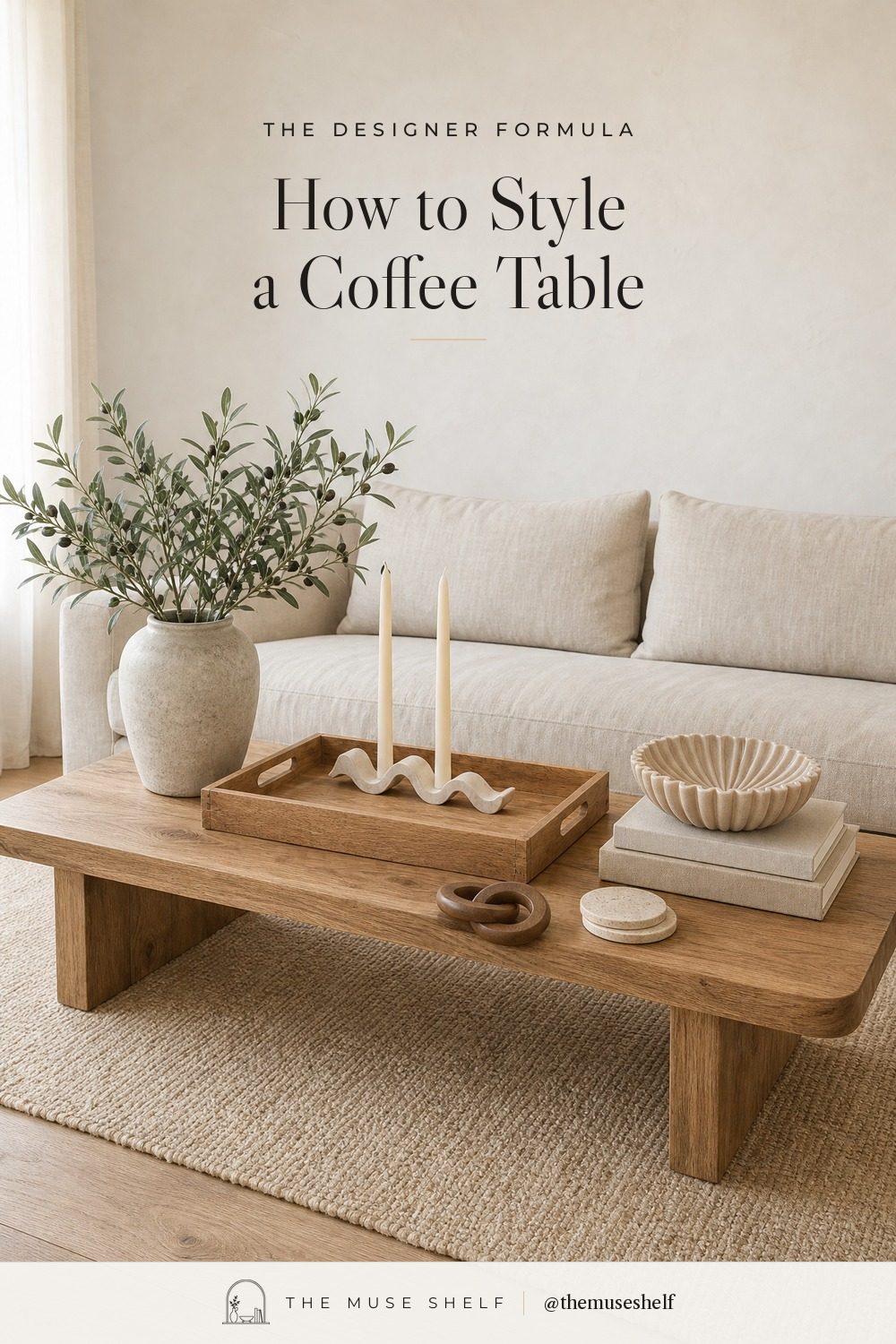 How to style a coffee table like a designer: a white oak coffee table in front of a cream linen sofa, styled with an acacia wood tray holding a white wave candle holder with ivory tapers and a wood chain link, a rustic cream vase of olive branches, a fluted bowl on two linen books and travertine coasters.