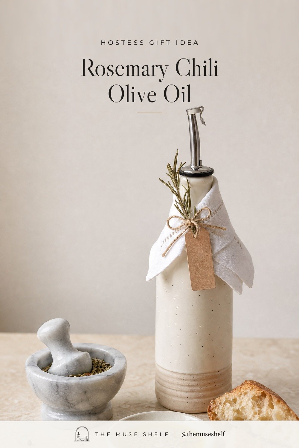 Cream speckled ceramic olive oil bottle with a steel pour spout, a white linen napkin tied at the neck with twine, a dried rosemary sprig and a blank kraft tag, next to a small marble mortar of crushed dried herbs and a dish of olive oil with bread