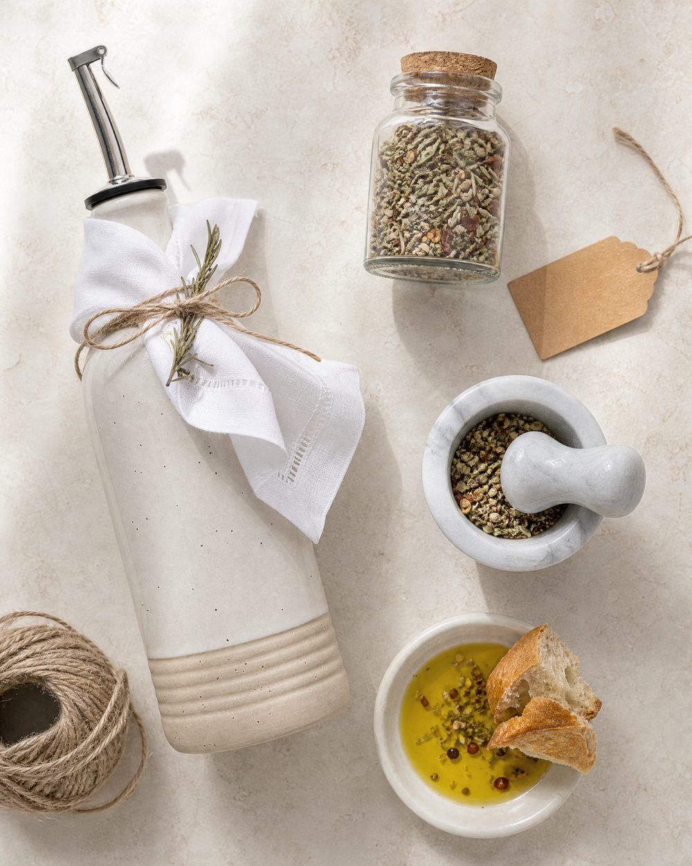 Overhead view of an olive oil gift kit: cream ceramic oil bottle wrapped with a linen napkin and twine, a cork-stoppered jar of dried herb and chili blend, a blank kraft tag, a marble mortar with the crushed blend and a dish of olive oil with bread